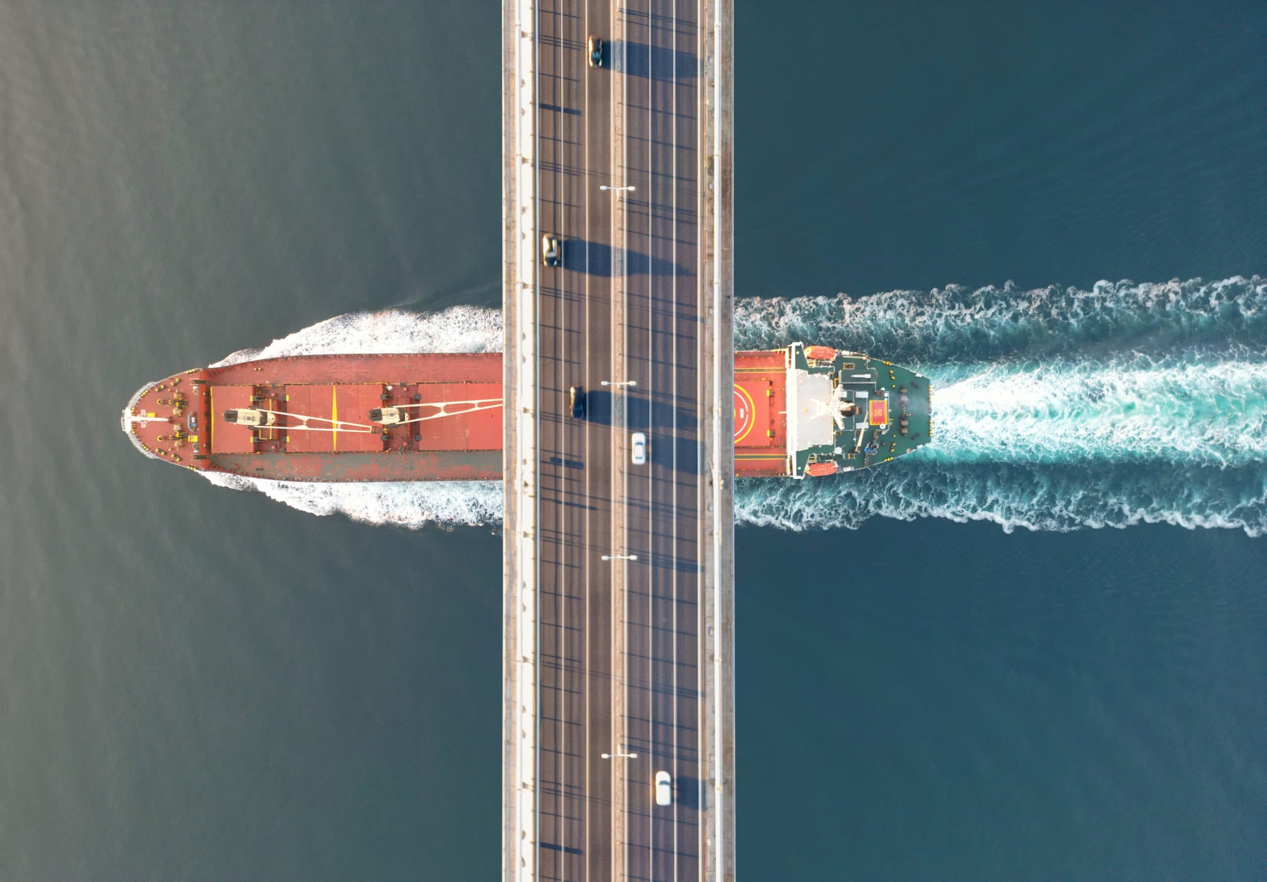 Aerial view illustrating the complex infrastructure of global trade, with a large cargo ship on an autonomous shipping route passing beneath a busy bridge, highlighting the advanced integration of logistics systems.